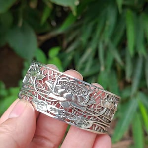 May include: Silver-toned cuff bracelet with a floral lotus design. The bracelet features intricate cut-out patterns and detailed lotus flower and leaf motifs. The metal bracelet is held against a blurred green background.