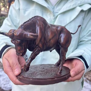 May include: A dark brown bull figurine standing in a charging pose on a circular base. The sculpture features detailed muscular anatomy, curved horns, and a thin tail extended outward, capturing a dynamic stance with one front leg slightly raised.