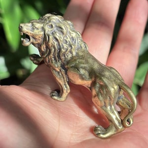 Könnte beinhalten: Kleine Löwenfigur aus Messing. Der Löwe wird brüllend dargestellt, mit offenem Maul und detaillierter Mähne. Die Figur hat ein gealtertes Aussehen mit sichtbarer Textur und Details.