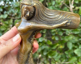 May include: Bronze mermaid door handle. The handle depicts a mermaid with flowing hair and a scaled tail. The mermaid's arm forms the handle. The bronze has a warm, antique finish. The handle is held by a hand.