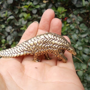 Könnte beinhalten: Kleine, detaillierte Pangolin-Figur aus Messing. Die Skulptur weist strukturierte Schuppen auf und wird in einer Hand vor einem Hintergrund aus grünem Laub gehalten. Das Pangolin ist ein einzigartiges Tier.