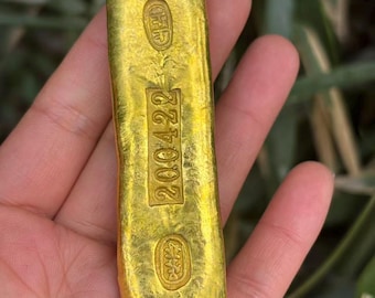 May include: A gold bar with embossed markings and the number "200422" stamped on it. The bar is rectangular with rounded edges and is held in a hand against a blurred green background.