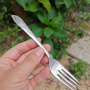 Op de afbeelding: Een zilveren vork met een bloemmotief op het handvat. De vork heeft drie tanden en wordt tegen een achtergrond van groen gebladerte gehouden. De vork lijkt vintage of antiek te zijn.