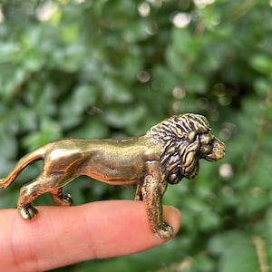 Könnte beinhalten: Kleine Löwenfigur aus Bronze. Der Löwe wird in einer Laufposition dargestellt, mit detaillierten Merkmalen wie einer strukturierten Mähne und muskulösen Beinen. Die Figur wird von einem Finger gehalten, mit einem verschwommenen grünen Hintergrund.