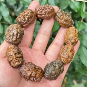 May include: A wooden bracelet featuring carved faces with intricate details. The bracelet is made of brown wood and is held in a hand against a backdrop of green foliage. The carved faces have varying expressions.