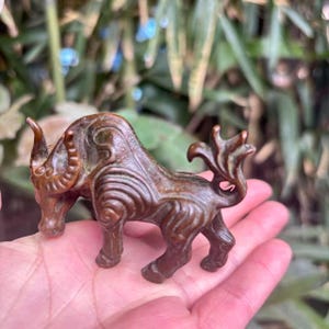 May include: A bronze bull figurine with intricate carvings. The bull features curved horns, a muscular physique, and a decorative tail. The figurine is held in a hand, with a blurred green backdrop.