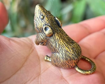 May include: A small, detailed brass mouse figurine. The mouse is depicted in a seated position with its tail curled. The brass has a textured surface and a warm, golden color. The figurine is likely a decorative item or collectible.