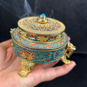 May include: An ornate teal and gold incense burner with a detailed floral design. The burner has a domed lid with a turquoise stone and is supported by three gold feet. Wisps of smoke rise from the top.