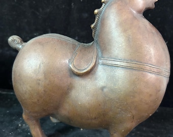 May include: A bronze-colored horse sculpture with a rounded body and a saddle. The horse has a detailed mane and tail, and is standing on four legs. The sculpture is likely made of metal.