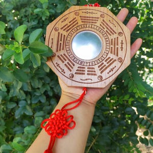 Peut inclure: Miroir Feng Shui octogonal en bois avec des symboles gravés et un miroir central circulaire. Le miroir est attaché à un nœud décoratif rouge. L'arrière-plan est un buisson vert luxuriant.