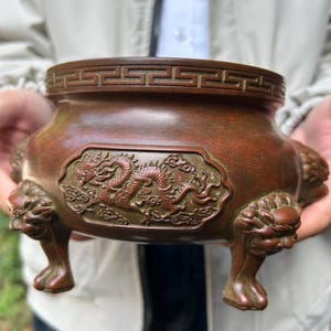 May include: A bronze incense burner with a detailed dragon relief. The vessel has a decorative rim and is supported by three lion-shaped feet. The overall color is a warm, reddish-brown.