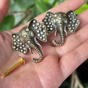 May include: Two antique brass elephant head cabinet knobs with detailed trunks and ears. A gold-colored screw is included. The knobs are likely for furniture or decorative purposes.