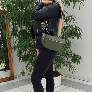 May include: A green leather fanny pack with a silver buckle and a long strap. The bag is worn over the shoulder of a person wearing a black leather jacket and black jeans.
