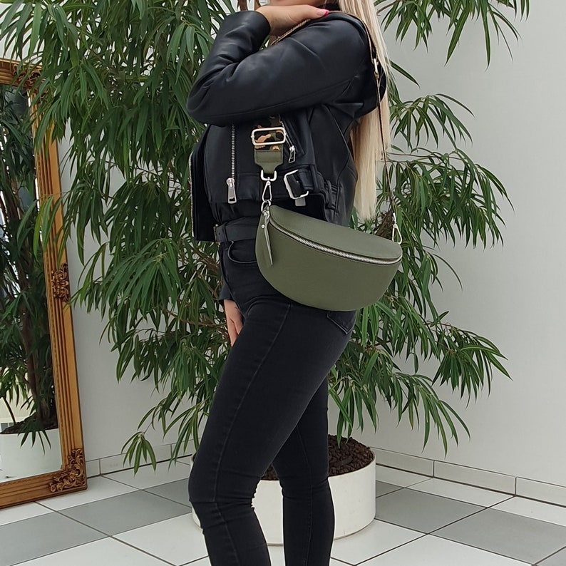 May include: A green leather fanny pack with a silver buckle and a long strap. The bag is worn over the shoulder of a person wearing a black leather jacket and black jeans.