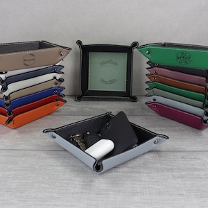 Könnte beinhalten: Ein Stapel von 10 Ledertabletts in verschiedenen Farben, darunter Orange, Rot, Blau, Beige und Weiß. Jedes Tablett hat einen genähten Rand und eine personalisierte Inschrift. Die Inschrift auf dem oberen Tablett lautet "The Weber Family". Das Tablett in der Mitte des Stapels hat ein grünes Innenleben und die Inschrift "Fischer Familie". Ein graues Tablett mit schwarzem Innenleben befindet sich im Vordergrund mit einem Telefon, Schlüsseln und einem kleinen weißen Gegenstand darin.