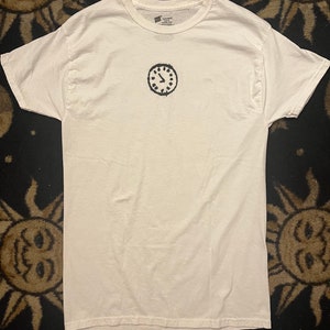 May include: White t-shirt with a black and white clock graphic on the front.