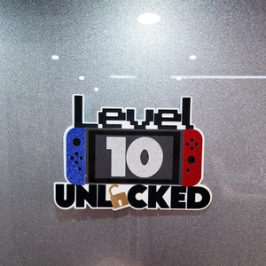 May include: A black and white graphic with the text "Level 10 Unlocked" in a pixelated font. The graphic features a blue and red Nintendo Switch controller with a silver number 10 on the screen.