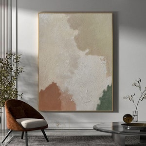 May include: Abstract painting with a textured background in shades of white, beige, brown, and green. The painting is framed in a gold frame and is hanging on a white wall.