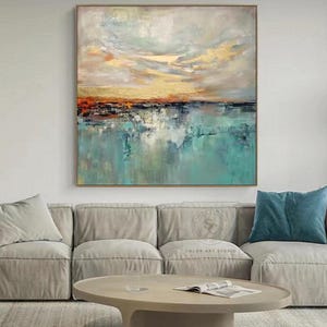 May include: Abstract landscape painting with a gold, orange, and teal color palette. The artwork features a textured sky and water reflection, framed in a gold-toned border. The painting is displayed above a gray sofa and a round coffee table.