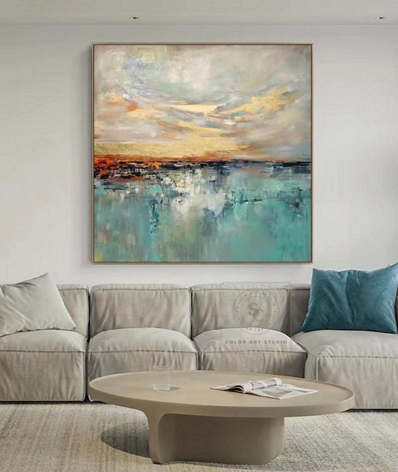May include: Abstract landscape painting with a gold, orange, and teal color palette. The artwork features a textured sky and water reflection, framed in a gold-toned border. The painting is displayed above a gray sofa and a round coffee table.