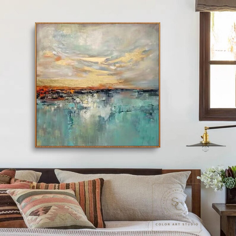 May include: Abstract landscape painting in a gold frame. The artwork features a sky with gold and orange hues above a turquoise and blue water reflection. The painting is a square format and hangs on a white wall.