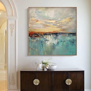 May include: Abstract landscape painting with a gold frame. The artwork features a serene scene with a turquoise body of water reflecting a colorful sky with orange and gold hues. The painting hangs above a dark wood cabinet.