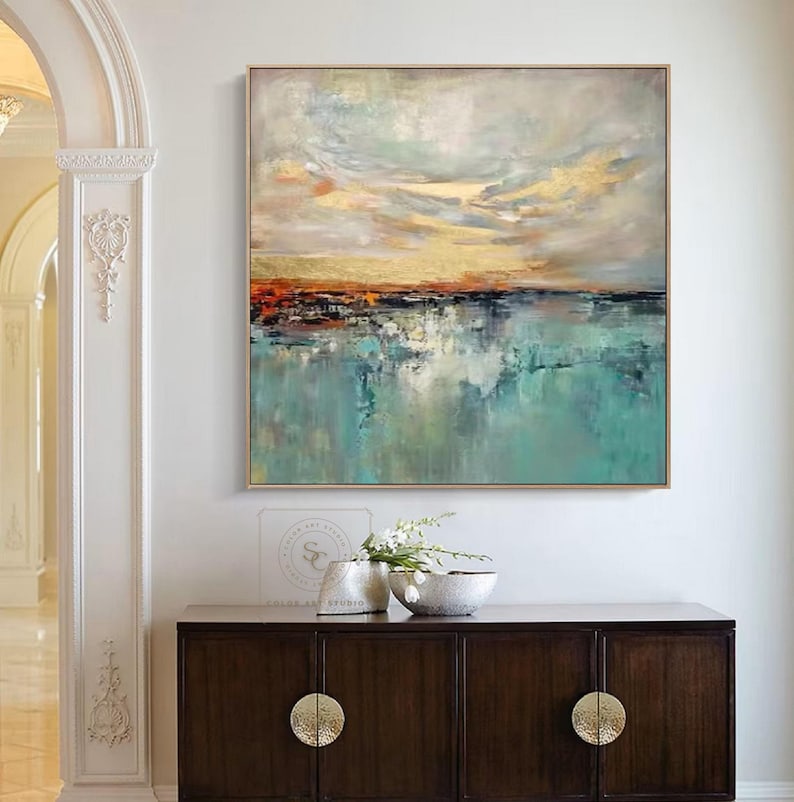 May include: Abstract landscape painting with a gold frame. The artwork features a serene scene with a turquoise body of water reflecting a colorful sky with orange and gold hues. The painting hangs above a dark wood cabinet.
