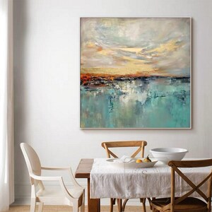 May include: Abstract landscape painting with a square silver frame. The artwork features a teal and blue water scene with a golden and orange sunset sky. The painting is displayed in a dining room setting.