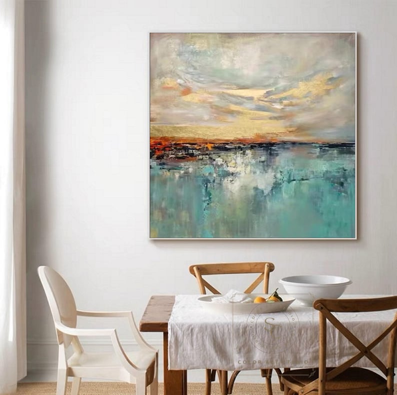 May include: Abstract landscape painting with a square silver frame. The artwork features a teal and blue water scene with a golden and orange sunset sky. The painting is displayed in a dining room setting.
