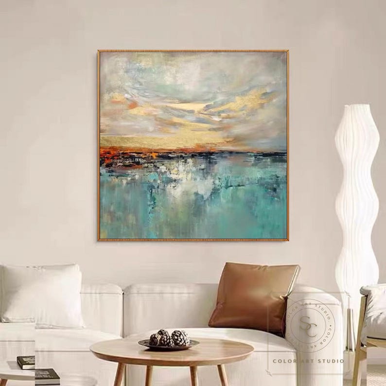 May include: Abstract landscape painting with a gold frame. The artwork features a serene scene with a turquoise body of water reflecting a cloudy sky with golden and orange hues. The painting is displayed above a white sofa.