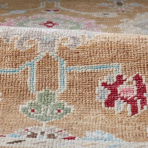 May include: Close-up of a rolled-up area rug with a detailed, woven pattern. The rug features a tan background with floral and geometric designs in red, pink, blue, and green. The texture of the rug is visible.