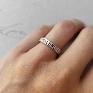 Könnte beinhalten: Ein silberner Ring mit dem Wort "WALDKIND" auf dem Band eingraviert. Der Ring wird an einem Finger vor einem weichen, neutralen Hintergrund gezeigt.