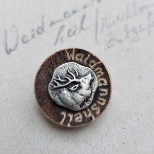 Könnte beinhalten: Runder, brauner Knopf mit einem silberfarbenen Hirsch-Emblem in der Mitte. Der Knopf trägt die Aufschrift "Waidmannsheil" am Rand. Der Hintergrund zeigt handschriftliche Schrift.