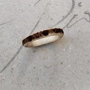 Könnte beinhalten: Ein handgefertigter Ring aus natürlichem Geweihmaterial. Der Ring hat eine hellfarbene Innenseite und eine dunkelbraune, strukturierte Außenseite. Der Ring wird auf einer neutralen, strukturierten Oberfläche gezeigt.