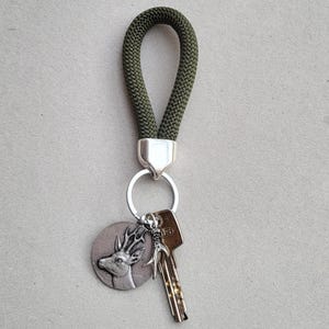 Könnte beinhalten: Ein Schlüsselanhänger mit einer dunkelgrünen Seilschlaufe und einem silberfarbenen Metallverschluss. Ein silberfarbener runder Anhänger mit einem Hirschdesign und ein Schlüssel sind am Schlüsselanhänger befestigt.