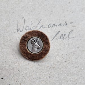 Könnte beinhalten: Ein runder, brauner Knopf mit einem silbernen Hirsch-Emblem in der Mitte. Der Knopf befindet sich vor einem strukturierten, hellfarbigen Hintergrund. Handschriftlicher Text ist im Hintergrund sichtbar.