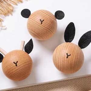 May include: Three wooden animal head ornaments. Each has a round, light brown wooden base with a black felt accent for the ears. The faces have a simple, dark brown 'Y' shape for the nose. One has round ears, one has long ears, and one has small ears.
