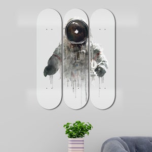 May include: Three white skateboard decks with a watercolor illustration of an astronaut in a spacesuit. The astronaut is wearing a helmet with a black visor. The illustration is in shades of gray and white.