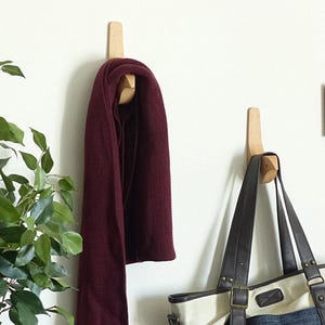 Könnte beinhalten: Zwei Wandhaken aus Holz mit einem einzigartigen geschwungenen Design. An einem Haken hängt ein burgunderfarbener Schal, am anderen eine braune und weiße Tragetasche.