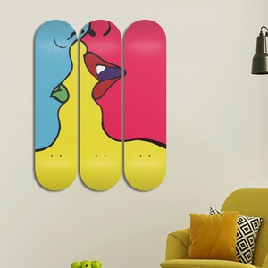 May include: Three skateboard decks with a colorful abstract painting of two faces kissing. The left face is blue, the middle face is yellow, and the right face is pink.