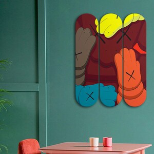Op de afbeelding: Drie kleurrijke skateboard decks met een cartoon-personage ontwerp. Het personage heeft bruine, rode en oranje huidtinten met geel haar en blauwe ogen. Het personage heeft een grote, rode neus en een paar doorgestreepte ogen. De decks hangen aan een turquoise muur.