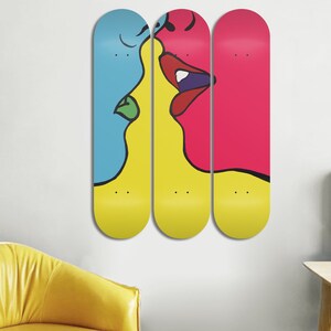 May include: Three yellow, blue, and pink skateboard decks with a black line drawing of two faces kissing. The faces are partially on each deck.