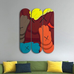 Op de afbeelding: Drie skateboard decks met een kleurrijk cartoonontwerp met een rode achtergrond, blauwe, gele, oranje en bruine handen en zwarte X'en.