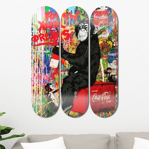 Op de afbeelding: Drie kleurrijke skateboarddecks met een spuitverfdesign. Het middelste deck heeft een zwarte chimpansee die op een rode Coca-Cola-koelbox zit. De tekst "Follow Your Dreams" staat op het linker deck.