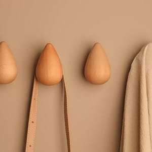 May include: Three wooden teardrop-shaped wall hooks in a light brown hue are mounted on a matching wall. A tan strap hangs from one hook, and a beige fabric hangs from the rightmost hook. The hooks are a minimalist design.