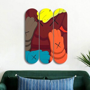 Op de afbeelding: Drie skateboard decks met een kleurrijk abstract ontwerp in een kleurenschema van rood, geel, blauw en oranje. Het ontwerp bevat een herhaald X-patroon en een cartoonachtige hand.