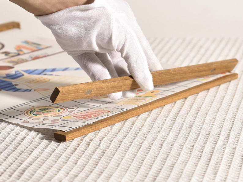 Puede incluir: Una mano con un guante blanco est&aacute; sujetando un marco de madera a una obra de arte impresa. La obra de arte est&aacute; sobre una superficie blanca texturizada.
