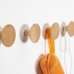 May include: A set of four wooden wall hooks with a natural finish. The hooks are circular with a cylindrical base, mounted on a white wall. An orange zippered pouch and white cords hang from two of the hooks.