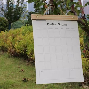 Puede incluir: Un planificador mensual blanco con una parte superior de madera, colgado al aire libre. El planificador tiene un diseño de cuadrícula para las entradas diarias y las palabras "Monthly Planner" en la parte superior. El fondo presenta follaje verde y un paisaje urbano.