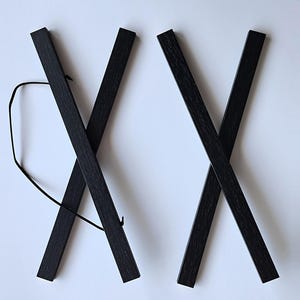 Pu&ograve; includere: Due portaposter in legno nero, ciascuno con un design a croce, sono mostrati su uno sfondo bianco. I portaposter sono collegati da uno spago nero.